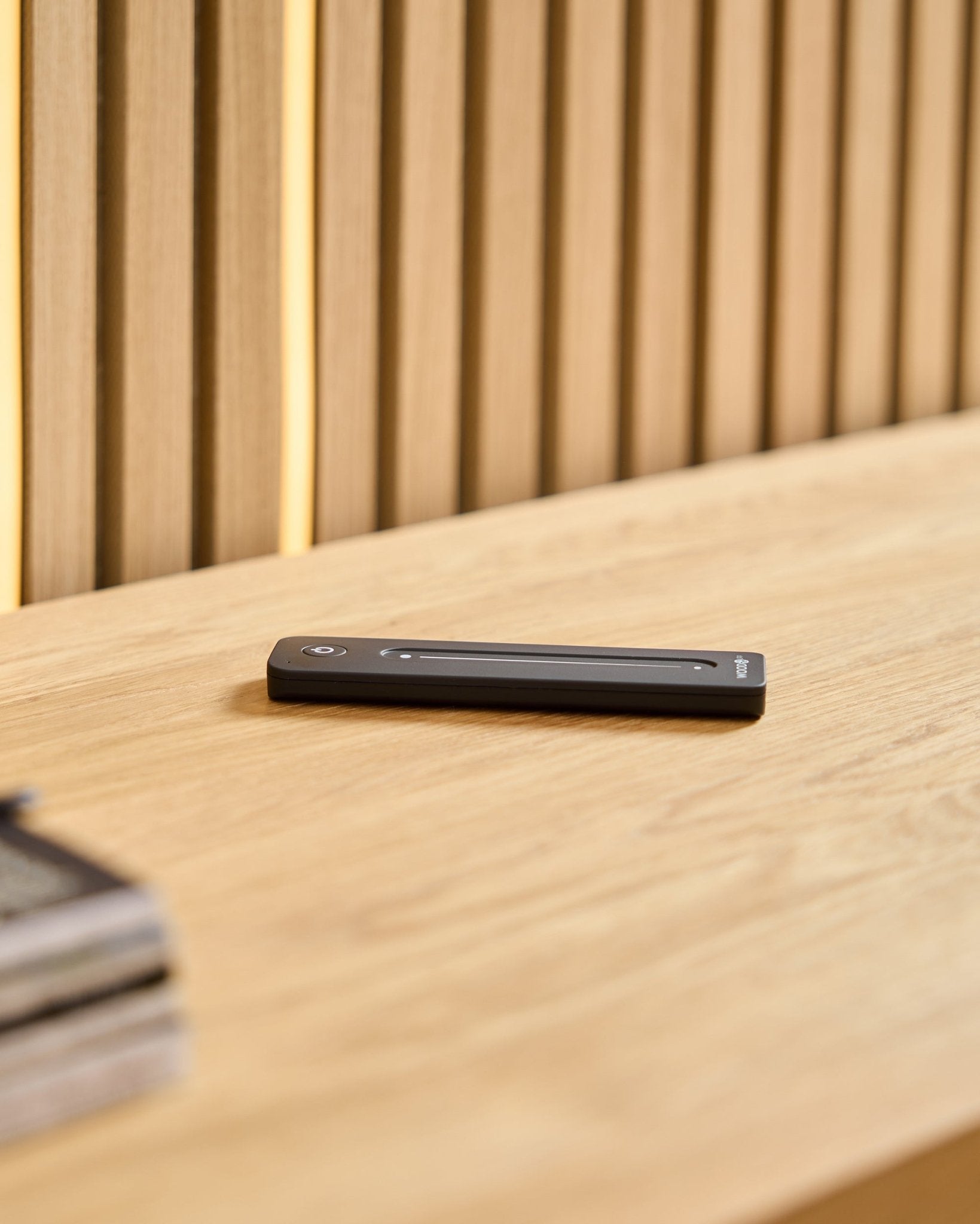 A black WoodUpp Lumi LED remote on a light wooden surface, set against a background of slatted acoustic wood panels.