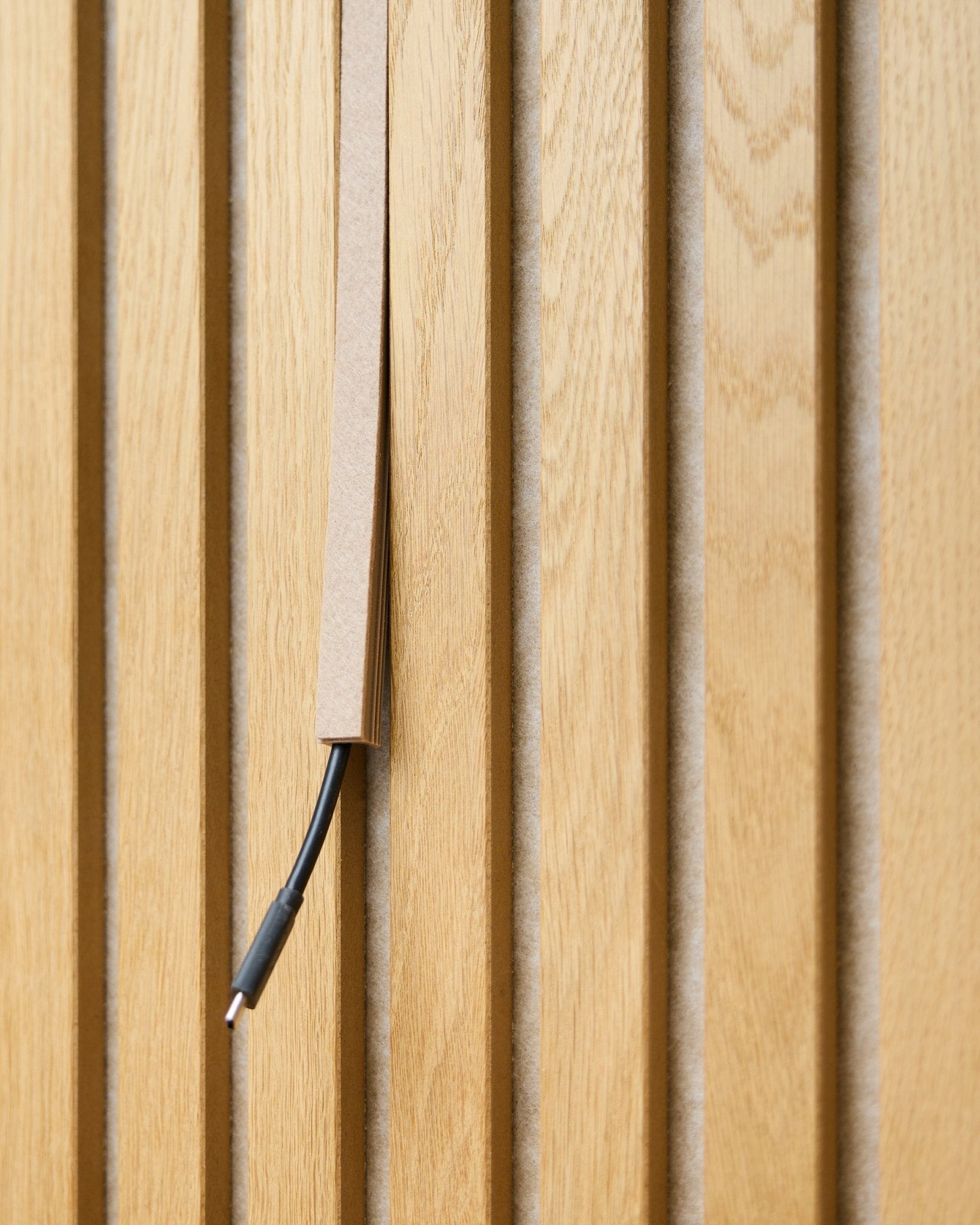 A black cable with cable management is partially concealed in a WoodUpp Dupps Beige Pop-on Cable Cover, mounted on vertical Akupanel acoustic wood slats.