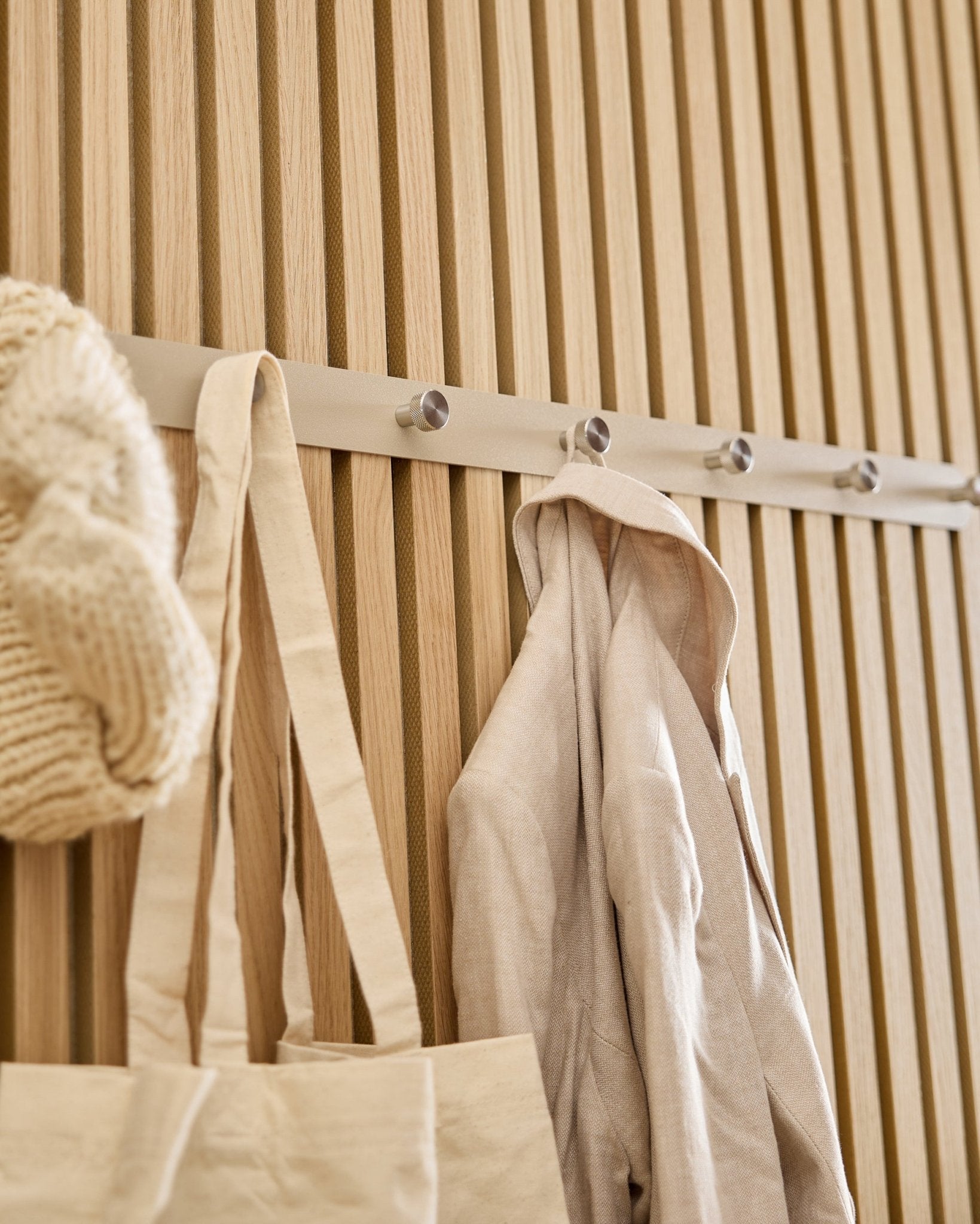 A beige tote, knit hat, and linen shirt hang on the WoodUpp Norr Coat Hook Rack (Beige/Stainless Steel, X7) mounted to a light wood Akupanel slatwall.