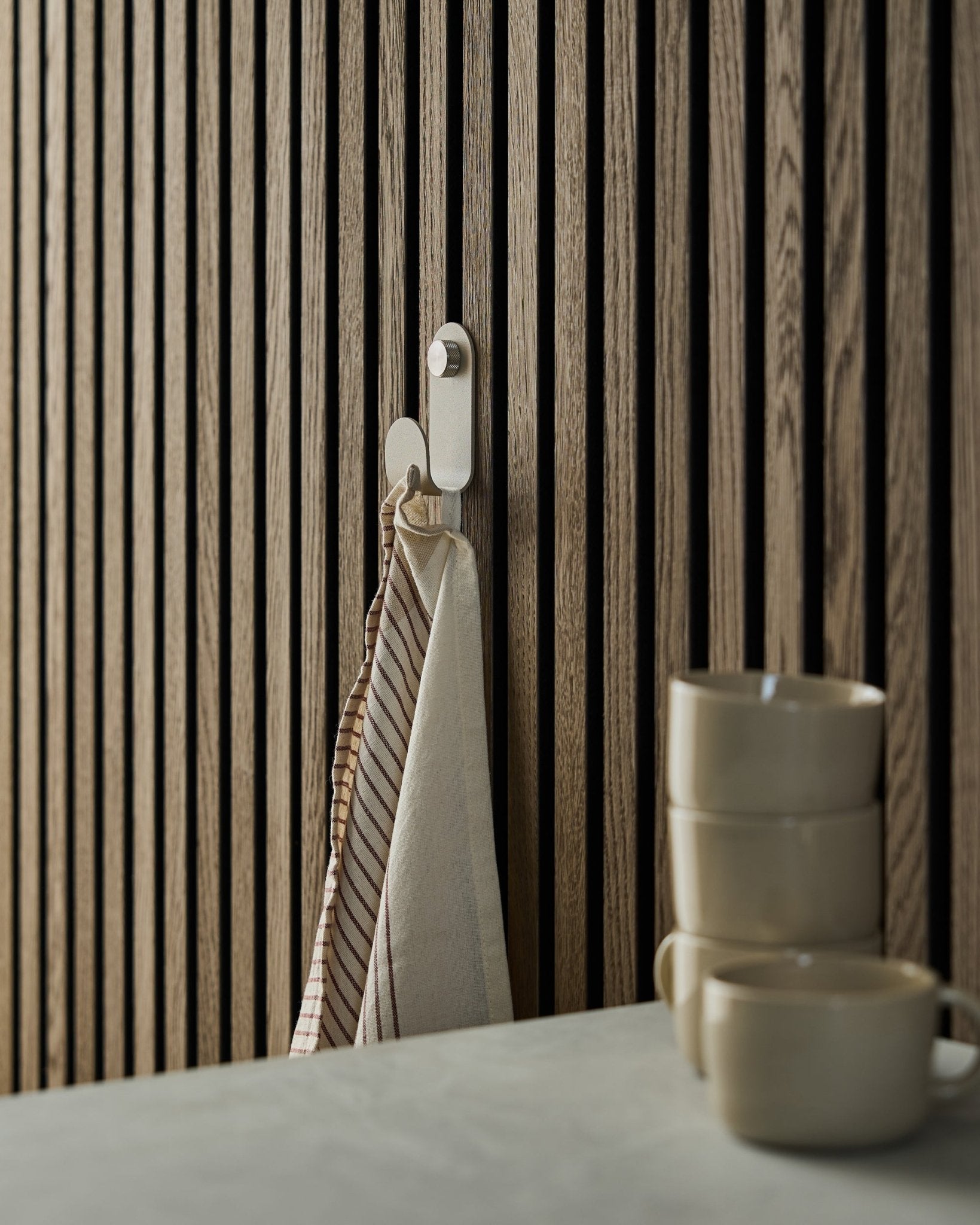 A striped towel hangs from a WoodUpp Norr Single Hanger in beige, mounted on a Nordic-style acoustic slatwall panel; three beige mugs are stacked in front.