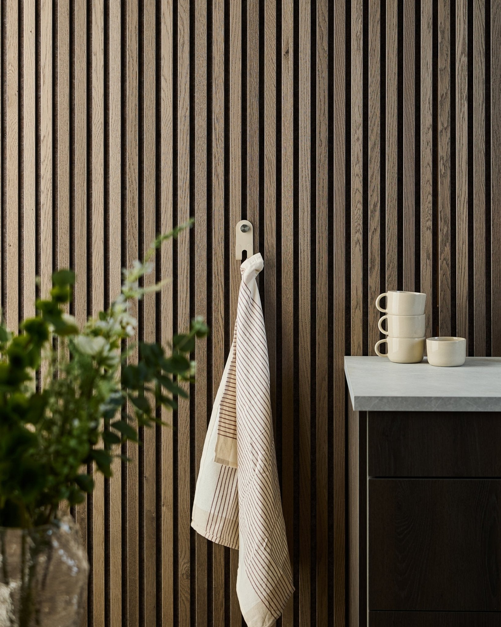 A striped towel hangs on the WoodUpp Norr Double Hanger in beige, mounted on a Nordic acoustic slatwall panel; mugs, bowl, and a green plant sit below.