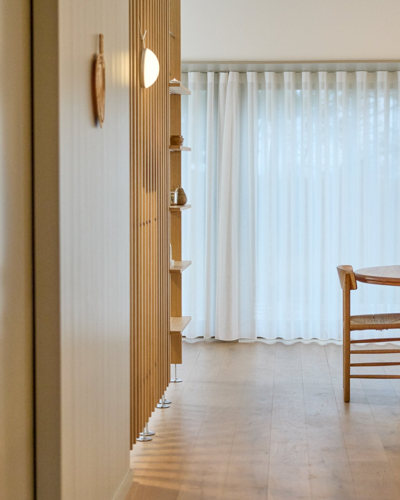 Minimalist room featuring a WoodUpp Embrace floor-to-ceiling room divider, wooden floor, shelves, chair, and sheer curtains over large windows.